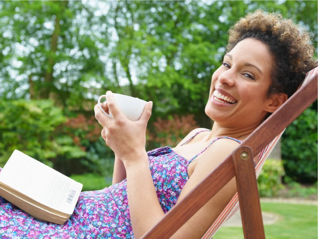 A woman enjoys a rest reset on a lounge chair outdoors, smiling as she holds a cup in one hand and a book in the other, surrounded by lush greenery.