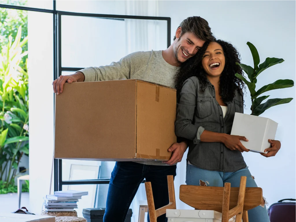 A man and woman stand indoors holding cardboard boxes and smiling, surrounded by moving boxes and houseplants.