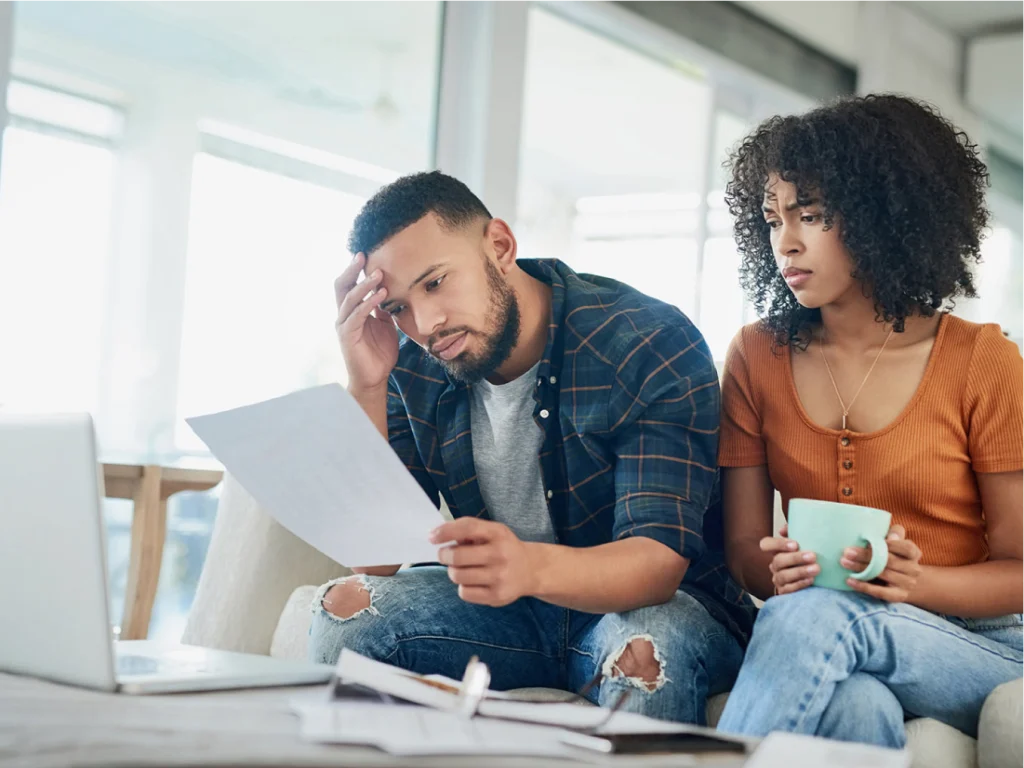 A man and woman sit on a couch looking concerned while reviewing papers in front of a laptop, possibly facing financial triggers; the woman holds a mug.