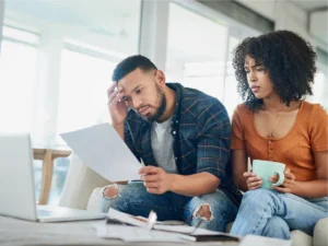 A man and woman sit on a couch looking concerned while reviewing papers in front of a laptop, possibly facing financial triggers; the woman holds a mug.