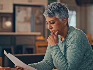 A woman with short gray hair wearing a light green sweater sits indoors, reading a piece of paper with a thoughtful expression, as if contemplating how to understand debt.