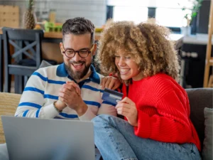 Two people sitting on a couch look at a laptop and smile, one holding a credit card, showing they're exploring how to spend your paycheck while shopping or making a purchase online.