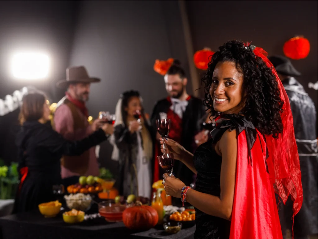 A woman in a red and black costume holds a glass of wine and smiles at the camera. Behind her, people in Halloween costumes gather around a table with snacks and drinks.