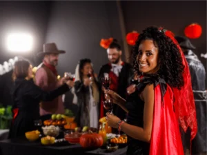 A woman in a red and black costume holds a glass of wine and smiles at the camera. Behind her, people in Halloween costumes gather around a table with snacks and drinks.