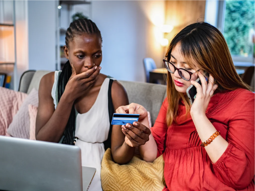 Two women sit on a couch; one is on the phone holding a credit card, while the other looks concerned with her hand over her mouth. A laptop is open in front of them.