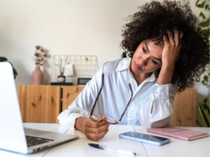 A woman sits at a desk holding her glasses and resting her head in her hand, appearing stressed, with a laptop, phone, notebook, and pen in front of her.
