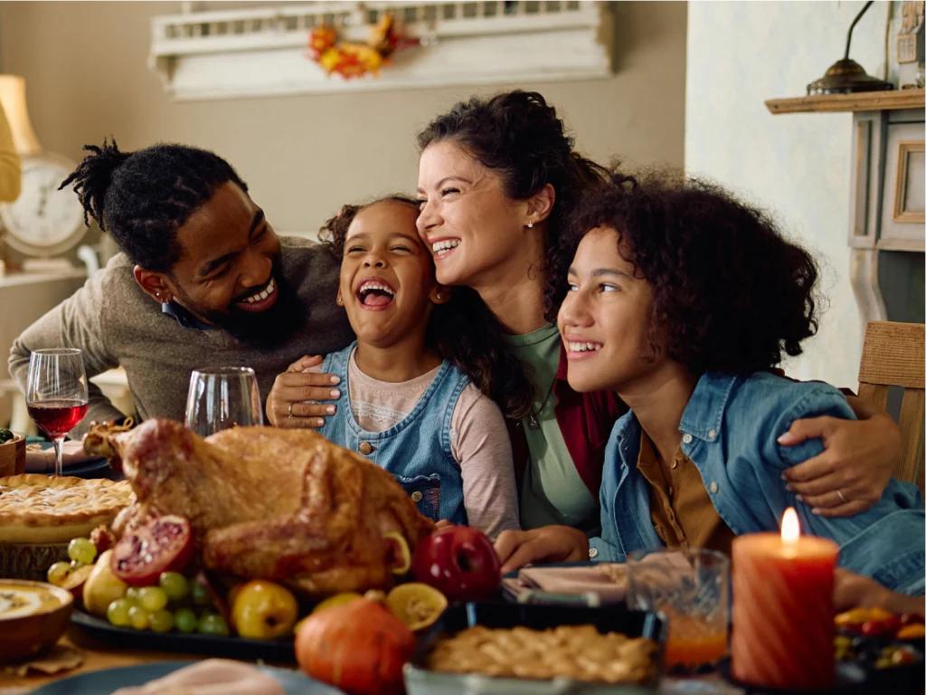 A family of four sits at a table with a roast turkey, pie, fruit, and candles, smiling and hugging during a festive meal.