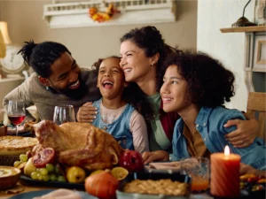 A family of four sits at a table with a roast turkey, pie, fruit, and candles, smiling and hugging during a festive meal.