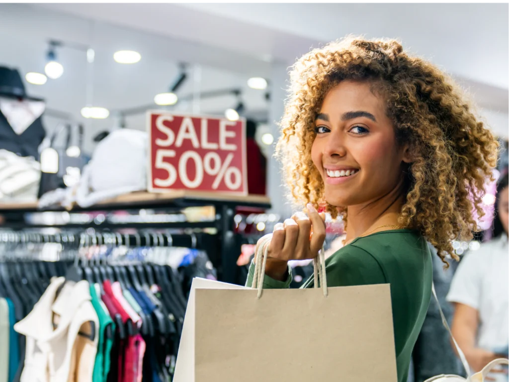 Woman holding a shopping bag smiles in a clothing store, with a 50% off sale sign visible in the background.