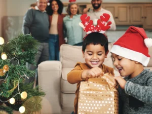 Two young children in festive hats open a present beside a decorated Christmas tree, with four adults smiling in the background.