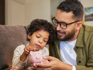 An adult and a child sit on a couch, with the child placing a coin into a pink piggy bank held by the adult.