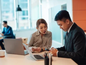 Two business professionals sit at a desk with laptops, discussing work while looking at documents, in a modern office setting.