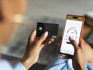 A person holds a black debit card and checks their credit score on a smartphone app, which displays a gauge indicating a good score.