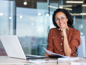 Woman with glasses in a brown polka dot blouse smiles at a desk with a laptop, holding papers and a pen in an office setting.