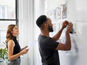 A man attaches a photo to a whiteboard while a woman holding papers watches and smiles in a bright office setting.
