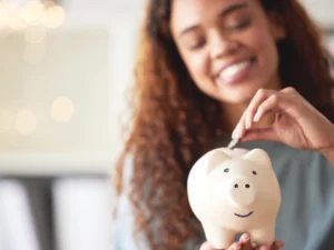 A woman smiles while putting a coin into a white piggy bank.