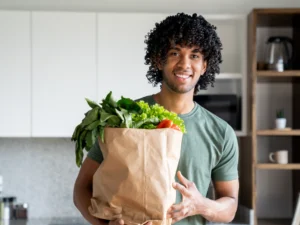 A person stands in a kitchen holding a paper bag filled with fresh vegetables and greens, smiling at the camera.