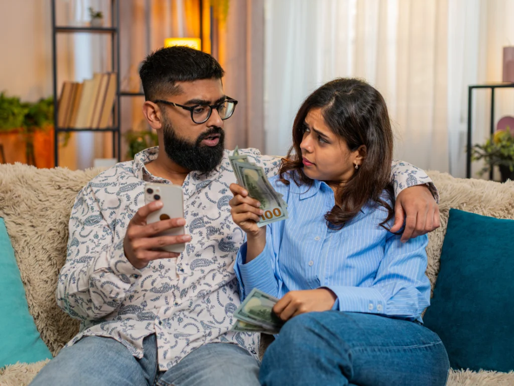 A couple sits on a couch; the man holds a phone and looks at the woman, who holds several $100 bills, as they appear to discuss money.