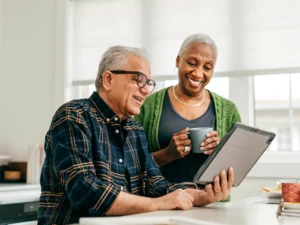 Two older adults sit at a kitchen counter, one holding a tablet and the other holding a mug, both smiling and looking at the tablet screen.