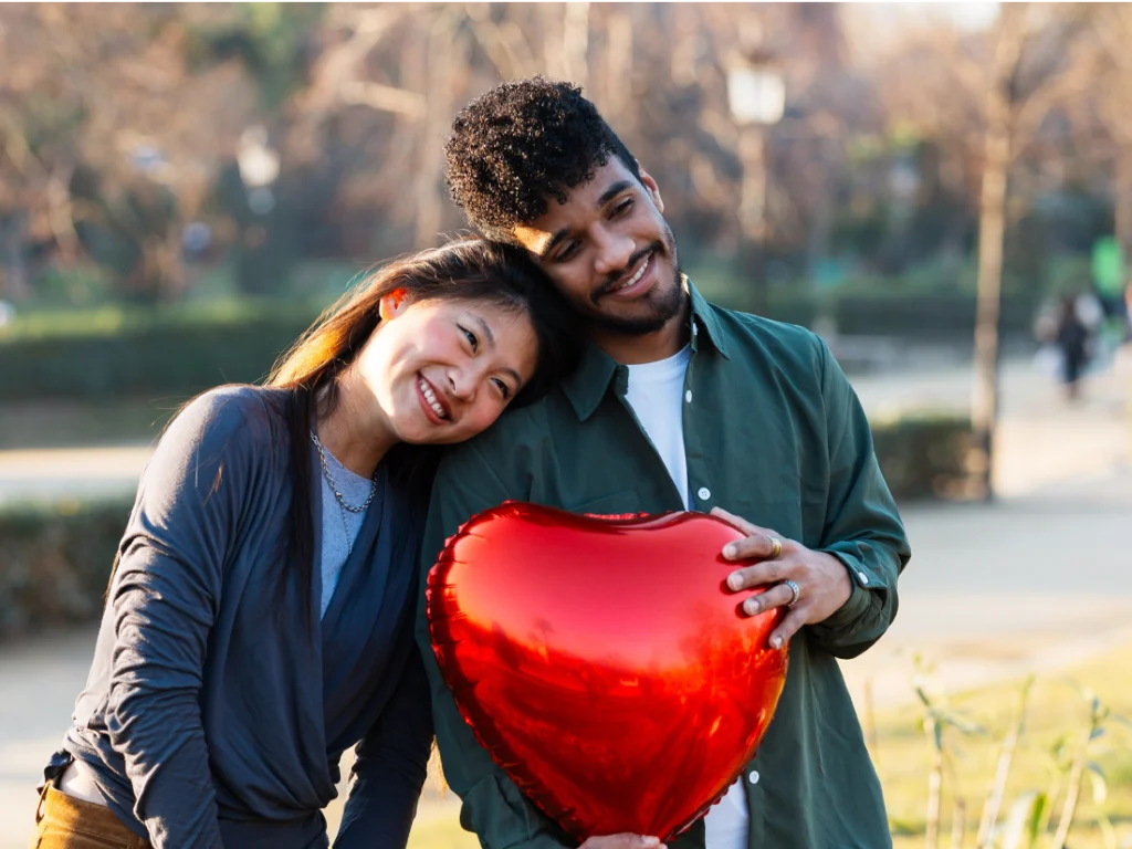 A woman and a man smile outdoors, leaning together and holding a large red heart-shaped balloon.