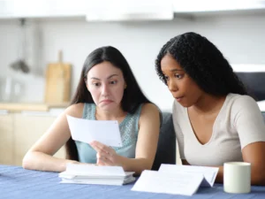 Two women sit at a table looking at documents with surprised and concerned expressions; a cup and a stack of papers are in front of them.
