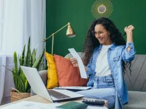 A woman with long curly hair sits on a couch, smiling and raising her fist while holding papers, with a laptop, notebook, and calculator on the table in front of her.