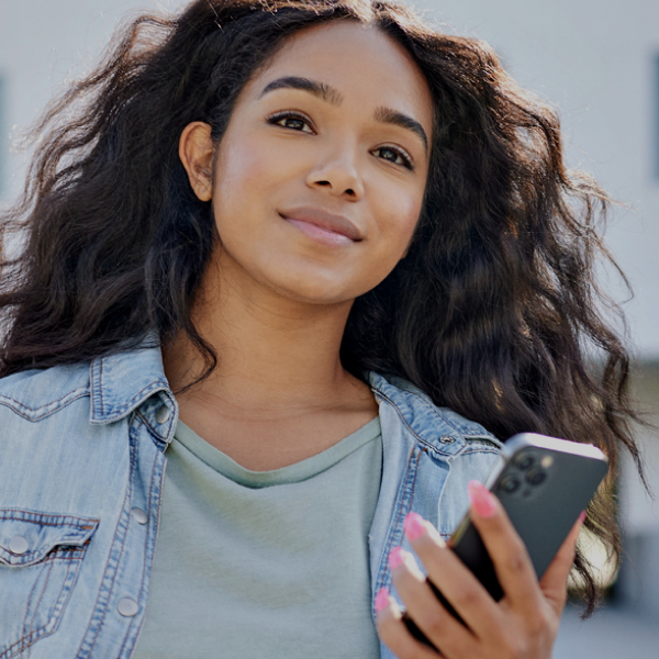 A woman with long, wavy hair holds a smartphone and smiles slightly while standing outdoors, wearing a denim jacket over a green shirt.