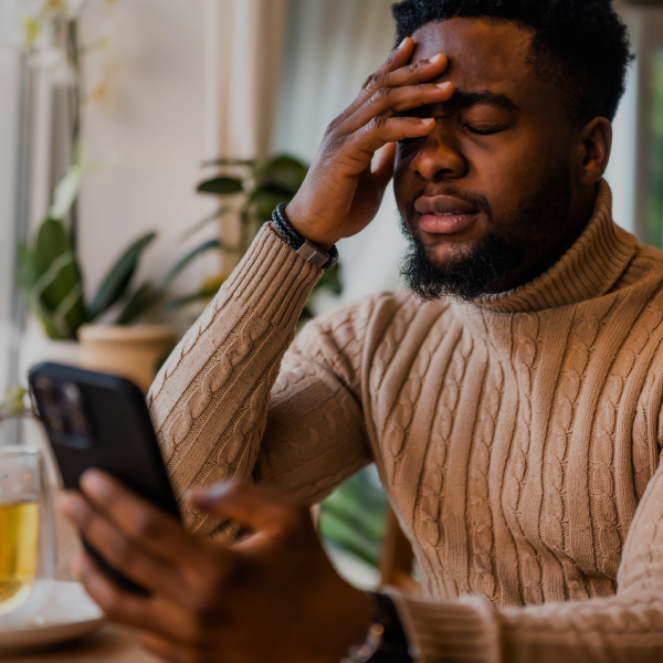 A man in a beige sweater sits at a table, holding a smartphone in one hand and covering his face with the other, appearing stressed or frustrated.