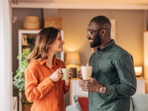 Two people stand indoors, facing each other and smiling, while holding mugs. A cozy living room with warm lighting is visible in the background.