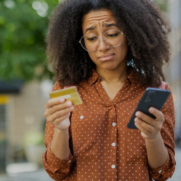 A woman with glasses looks at a credit card and a smartphone with a concerned expression, standing outdoors in a brown polka-dot shirt.