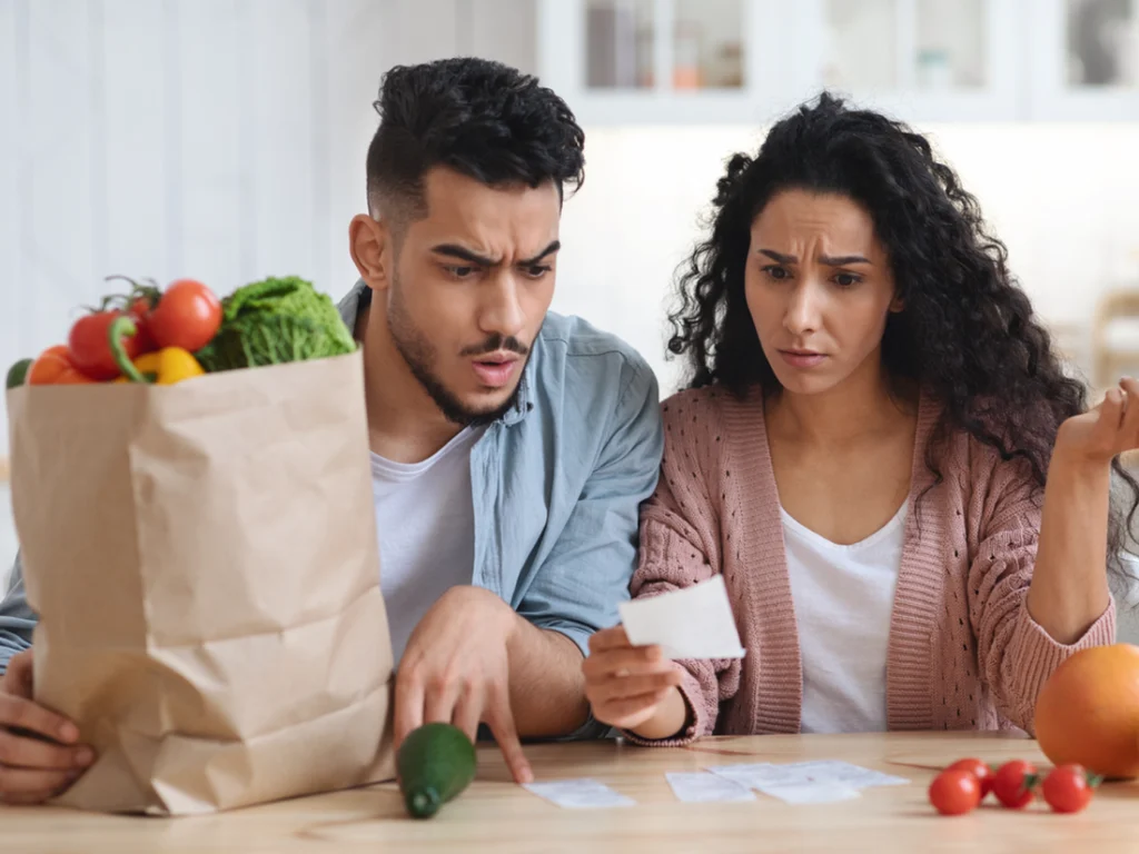 A man and woman sit at a table with a grocery bag and vegetables, looking concerned as they review a receipt.