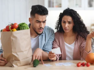 A man and woman sit at a table with a grocery bag and vegetables, looking concerned as they review a receipt.