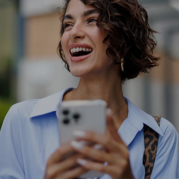 A woman with short curly hair smiles and holds a smartphone outdoors, wearing a light blue shirt and gold hoop earrings.