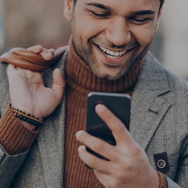 Man wearing a gray blazer and brown turtleneck smiles while looking at his smartphone, holding a bag strap over his shoulder.