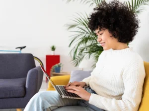 Person with curly hair sits on a yellow chair, smiling while using a laptop. Background includes a gray chair, houseplants, and a bicycle wheel.
