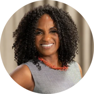 Woman with curly black hair, wearing a gray sleeveless top and a red beaded necklace, smiling in front of a neutral background.