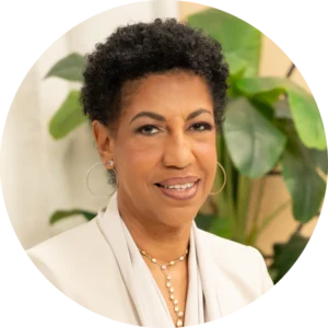 A woman with short curly hair wearing a light-colored blazer and necklace smiles in front of green indoor plants.