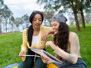 Two young women sitting on the grass outdoors, looking at and discussing a sketchbook together.