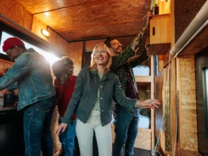 Four people are inside a small, wood-paneled room, smiling and engaged in different activities, including adjusting something on the wall and preparing food.