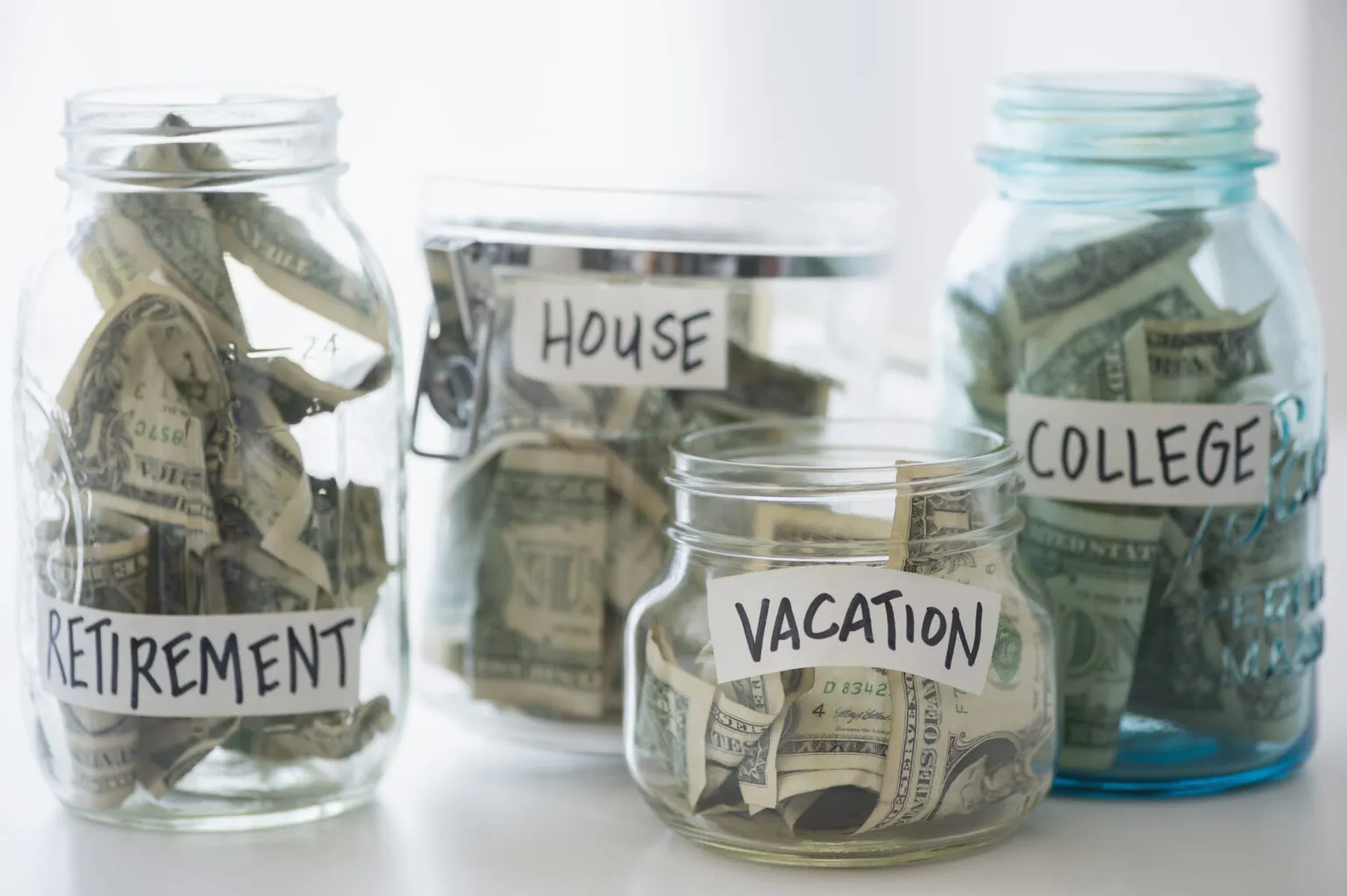 Four glass jars labeled retirement, house, vacation, and college, each filled with U.S. dollar bills, representing different savings goals.