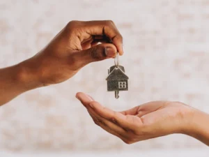 Two hands exchanging a house-shaped keychain, symbolizing the handover or receiving of house keys.