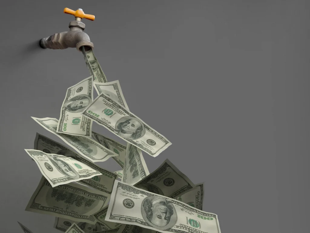 Dollar bills flowing out of a metal water faucet with a yellow handle against a gray background.