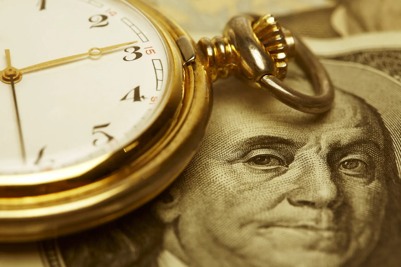 A gold pocket watch rests on top of a U.S. hundred-dollar bill, showing part of Benjamin Franklin's face.