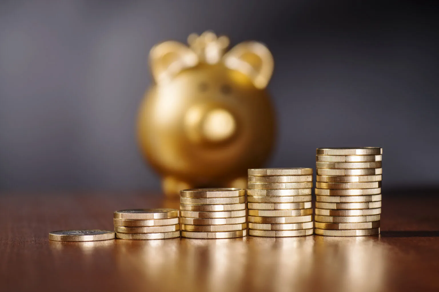 Stacks of gold coins are arranged in increasing height on a wooden surface, with a blurred golden piggy bank in the background.