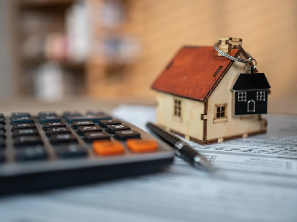 A calculator, a pen, and a miniature house keychain are placed on top of paperwork, representing real estate or mortgage concepts.