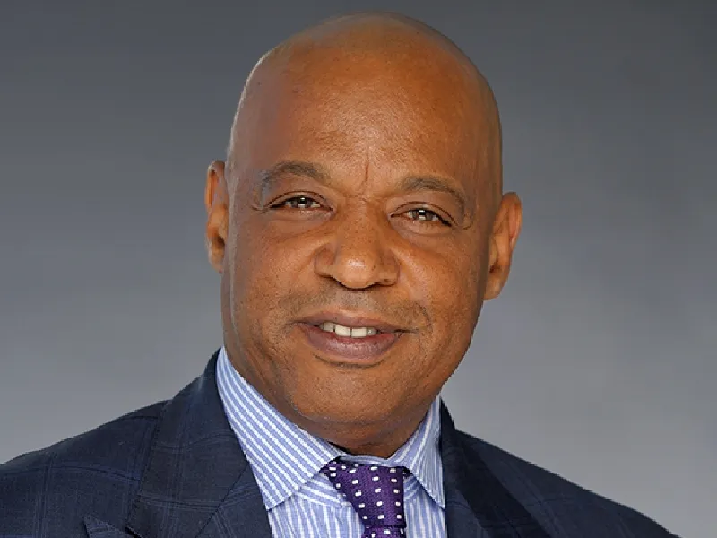 A middle-aged man wearing a dark suit, striped dress shirt, and purple polka dot tie, smiling at the camera against a plain gray background.
