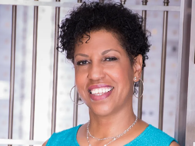 A woman with short curly hair, wearing hoop earrings and a blue sleeveless top, smiles in front of a metallic background with vertical bars.