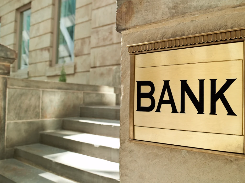 A brass sign with the word "BANK" mounted on a stone building beside concrete steps.