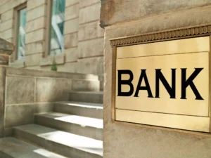 A brass sign with the word "BANK" mounted on a stone building beside concrete steps.