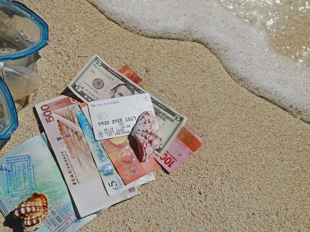 Various international banknotes, seashells, and a credit card are placed on sandy beach near the water's edge, with snorkeling gear partially visible on the left.